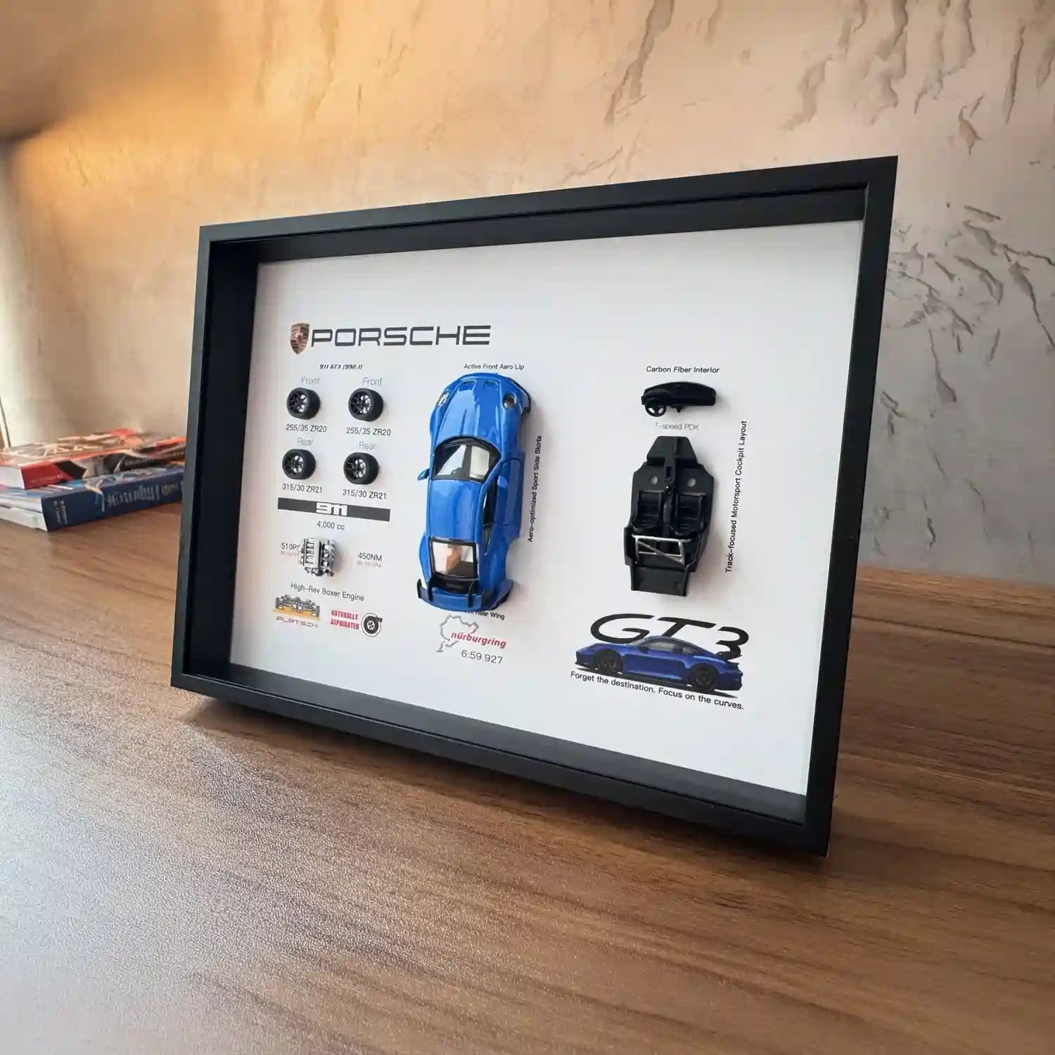 Porsche 911 GT3 992 Shark Blue Deconstructed Frame — angled on a wood desk against a textured plaster wall, books to the left